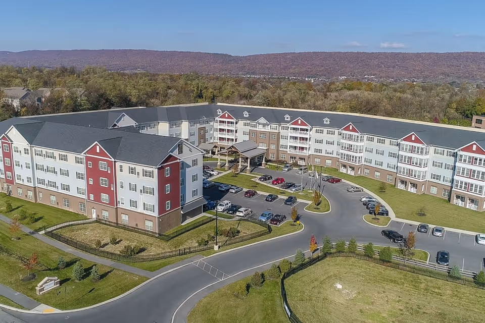 Aerial view of a large senior living facility named Harmony at West Shore, featuring a multi-story building with red and white exterior walls, a parking lot with several cars, surrounding greenery, and a backdrop of forested hills under a clear blue sky.