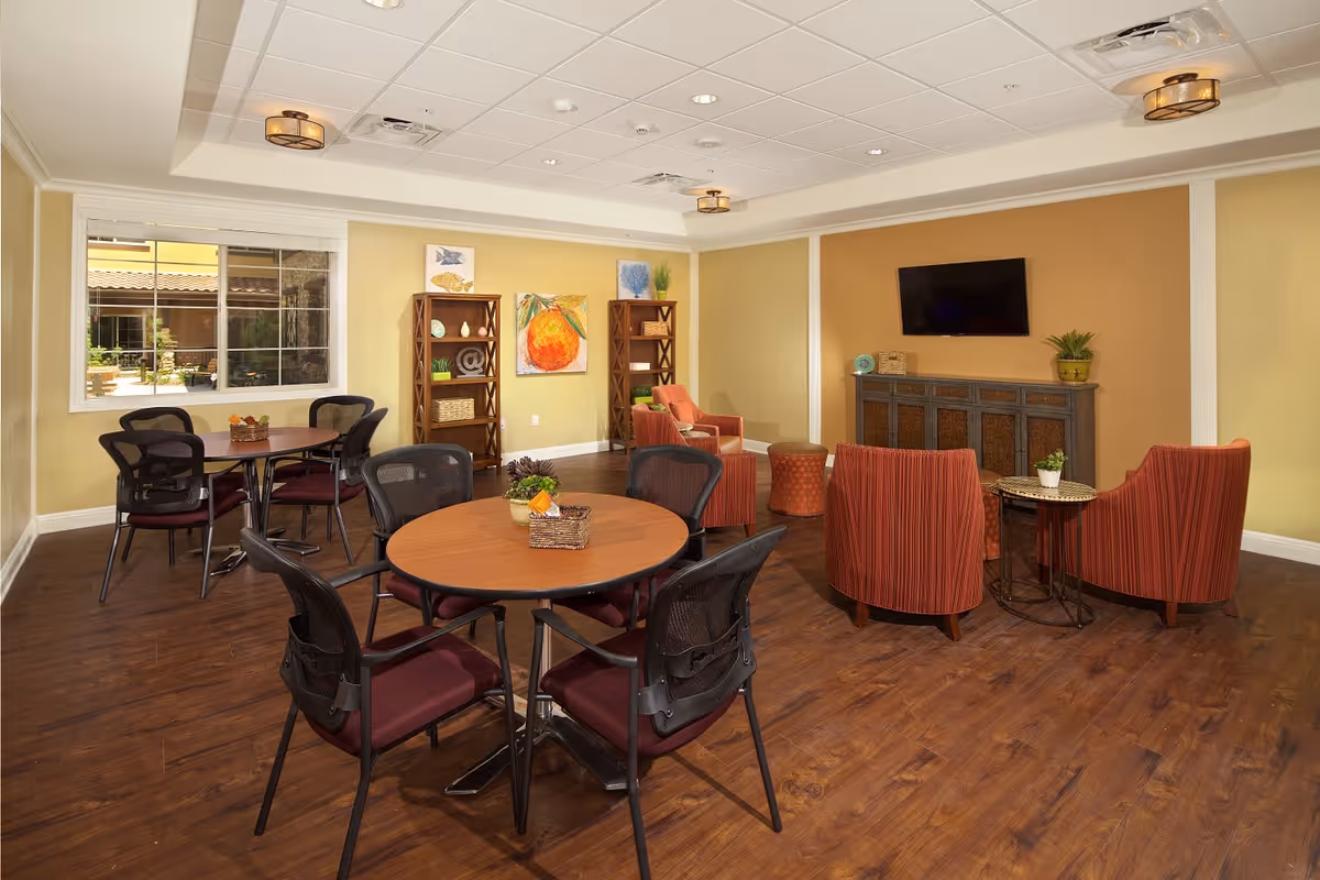 A cozy common area with round tables and chairs, a seating area with four red armchairs around a small table, a wall-mounted TV above a cabinet, and two wooden shelves with decorative items. The room has wood flooring, yellow walls, and a window showing an outdoor view.