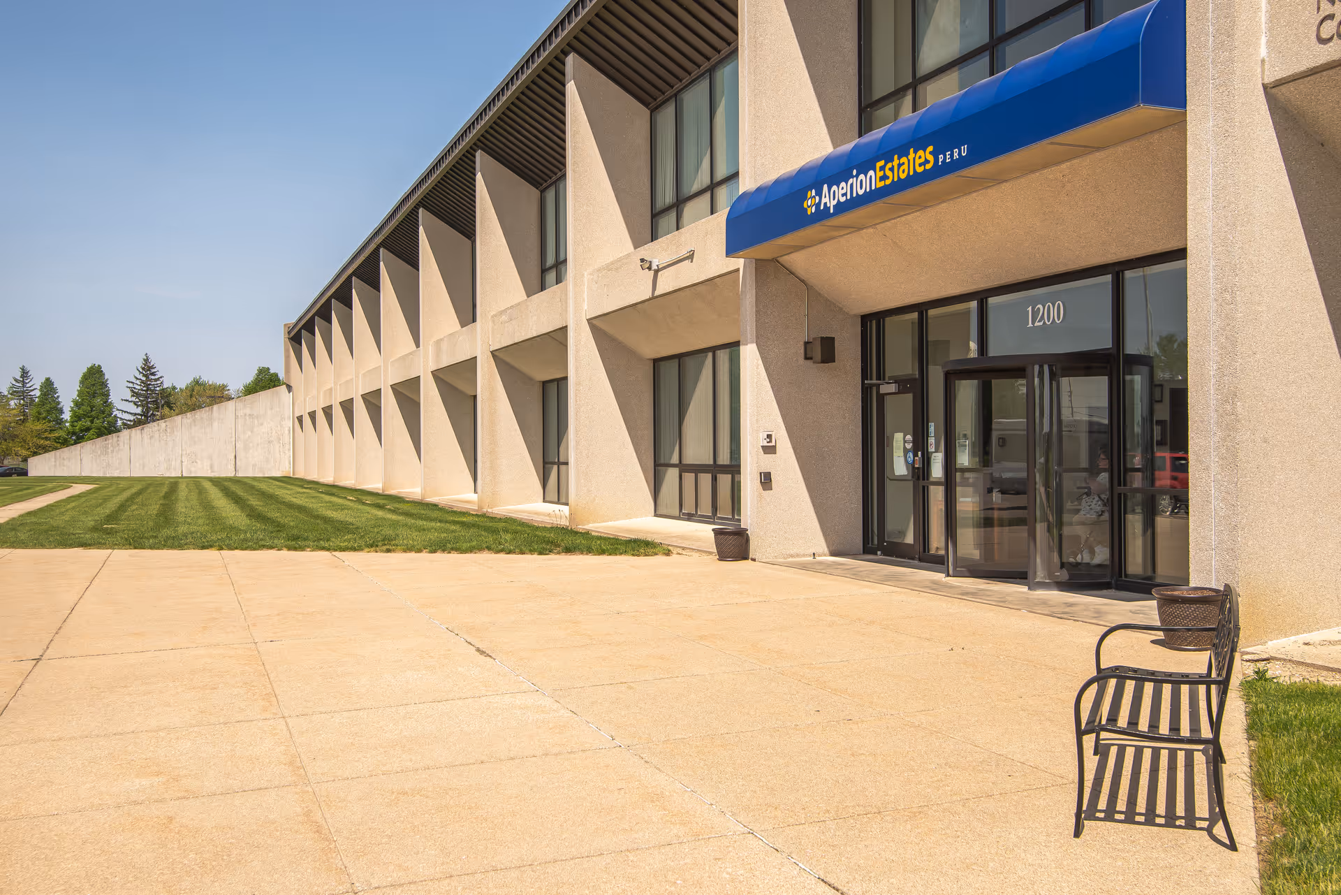 Exterior view of Aperion Estates Peru building with a blue awning above the entrance door. The building has large windows and a concrete walkway with a black metal bench on the right side. There is a well-maintained grassy area to the left of the walkway.