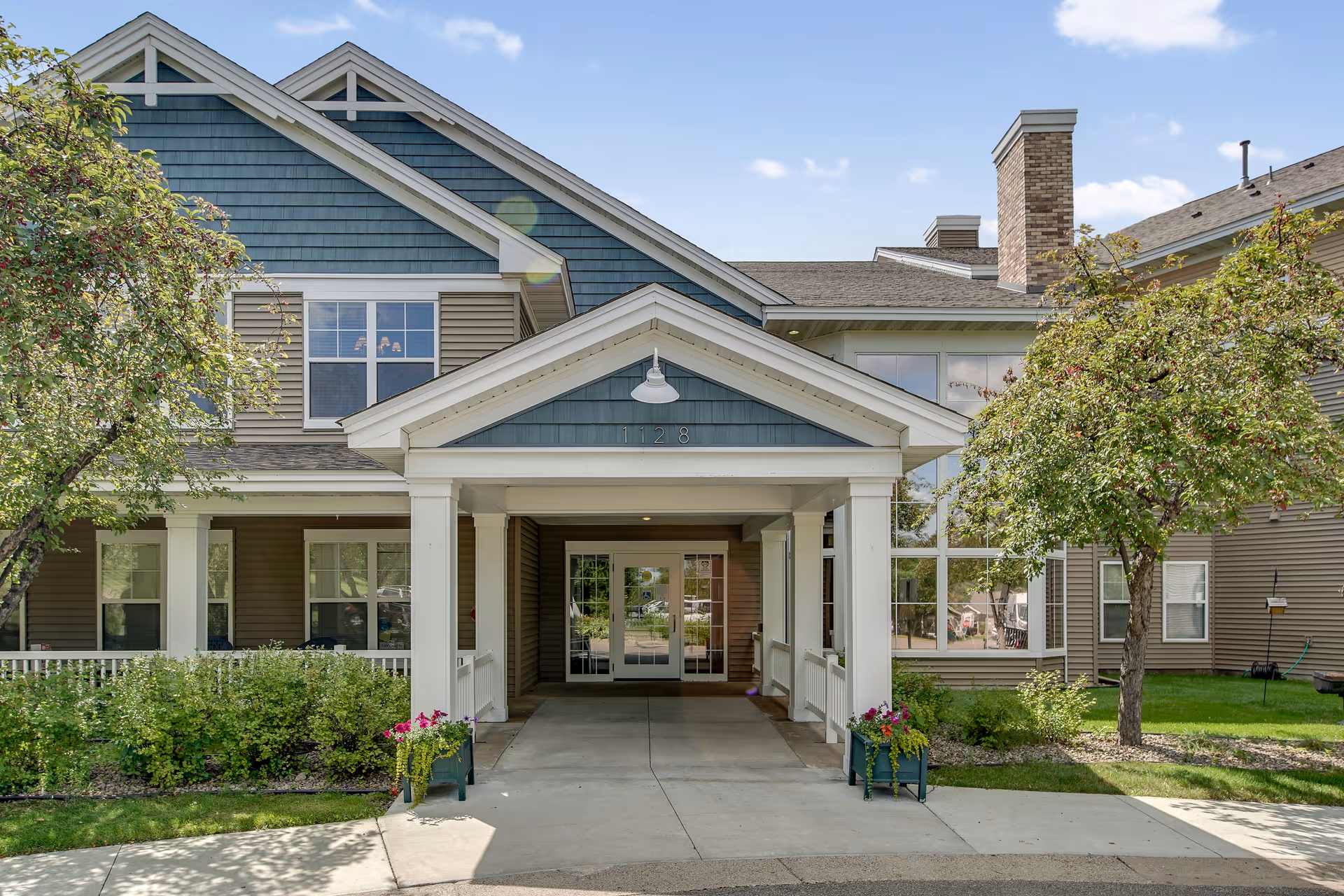 Front exterior view of Oak Ridge Assisted Living facility showing a covered entrance with white pillars, blue and beige siding, windows, and landscaped greenery with trees and flowers on either side of the walkway.