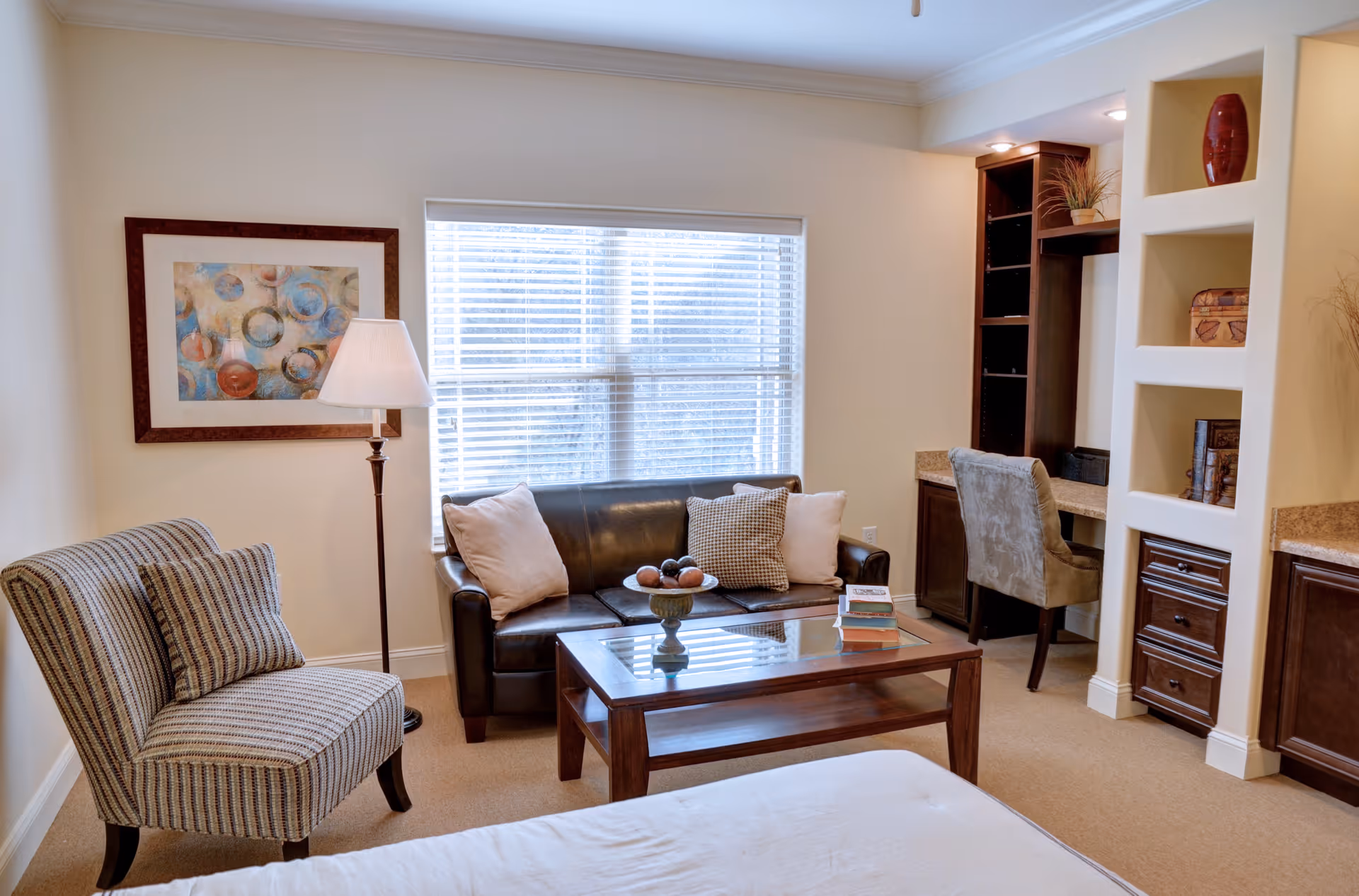 A cozy living area with a dark leather sofa adorned with pillows, a striped armchair, a glass-top wooden coffee table with decorative items and books, a floor lamp, a framed abstract painting on the wall, and a built-in desk with a chair and shelves. The room has beige walls and carpet, and a large window with blinds letting in natural light.
