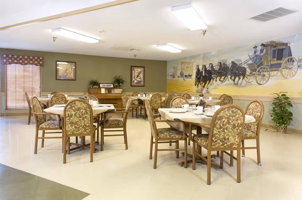 Dining room with round tables and patterned cushioned chairs set for meals and a large mural of a stagecoach on the wall.
