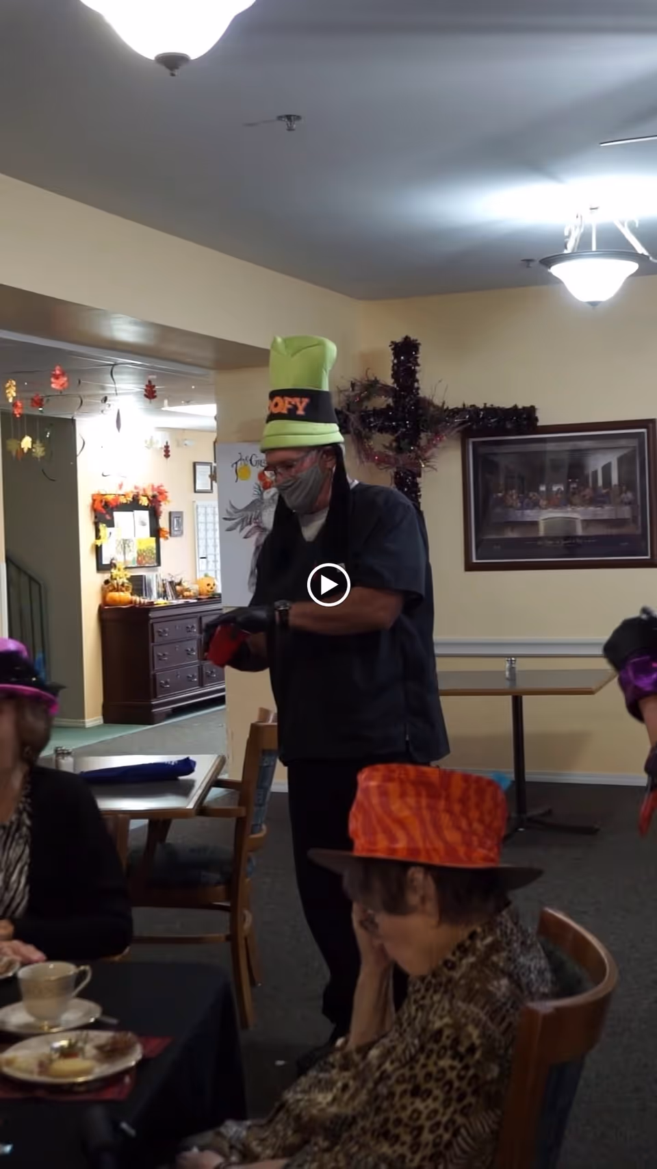 A group of elderly people wearing colorful hats are gathered in a decorated indoor room. One man in a green tall hat and face mask stands near a table, while two seated women wear orange and purple hats. The room has autumn-themed decorations, a framed picture on the wall, and tables with chairs.