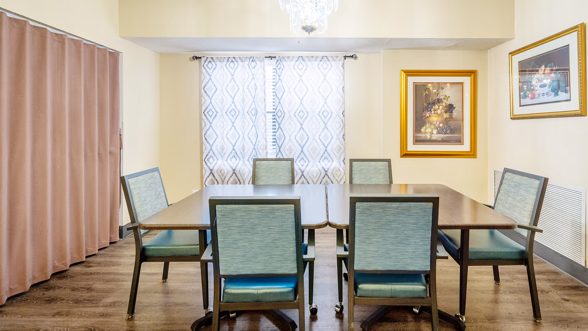 A dining room with a rectangular wooden table surrounded by six green cushioned chairs. The room has wooden flooring, beige walls, two framed paintings on the right wall, and a window with patterned curtains at the back. A chandelier hangs from the ceiling.