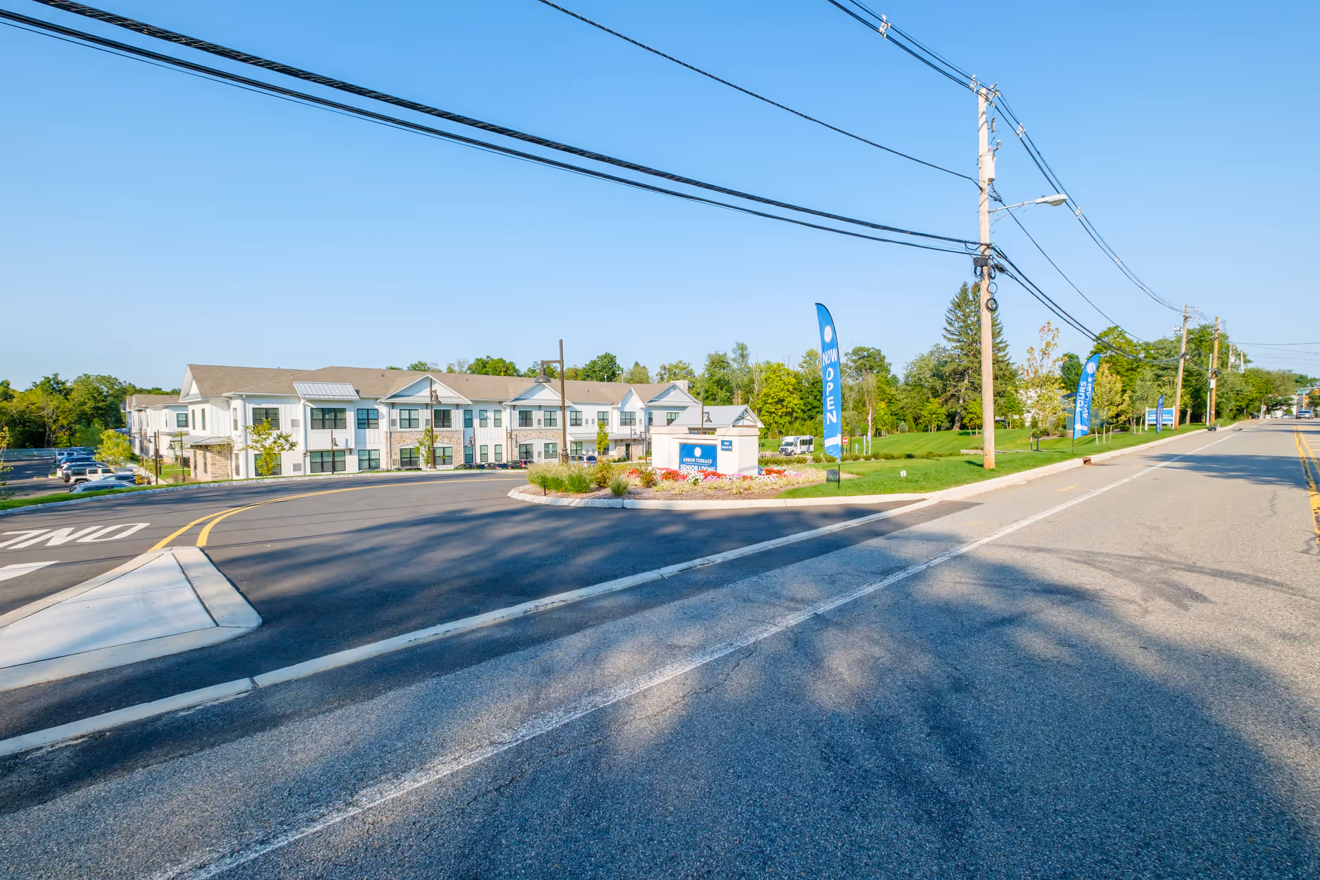 Exterior view of Arbor Terrace Basking Ridge senior living facility on a clear sunny day, showing a two-story building with a parking lot to the left and a landscaped area with flags that say 'NOW OPEN' and 'TOURS AVAILABLE' near the entrance.