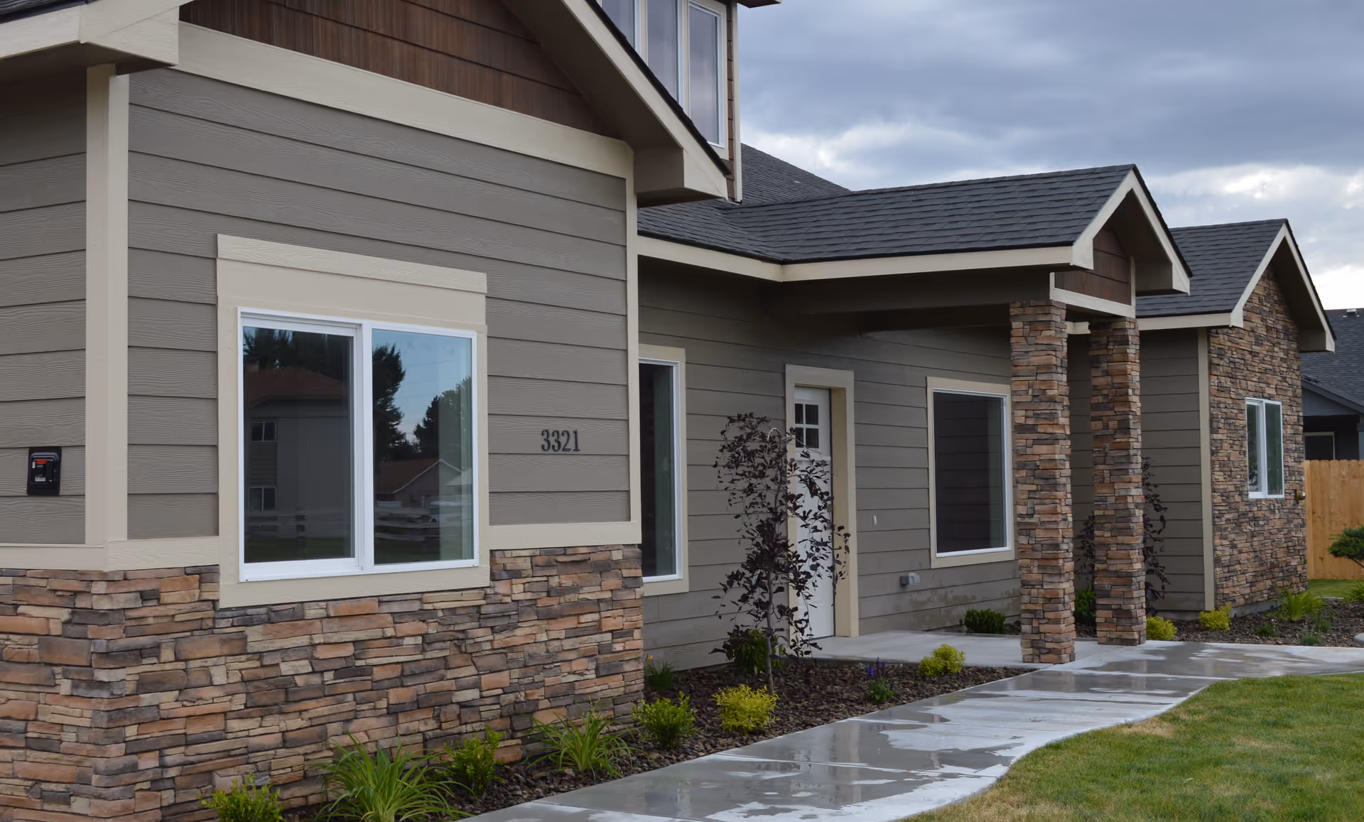 Exterior view of a modern senior living facility building with gray siding and stone accents. The building has multiple windows, a white door, and a covered entrance supported by stone pillars. The sidewalk and lawn in front are wet, indicating recent rain.