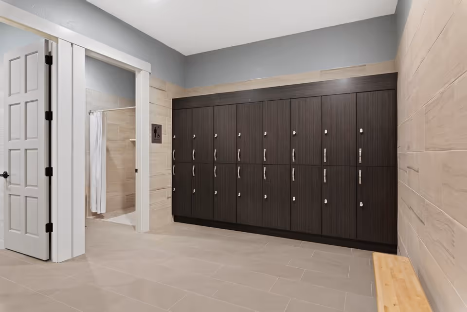 A clean locker room with a row of dark wood lockers along the wall, a light wood bench on the right, beige tiled floor and walls, and an open door leading to a shower area with a white curtain.