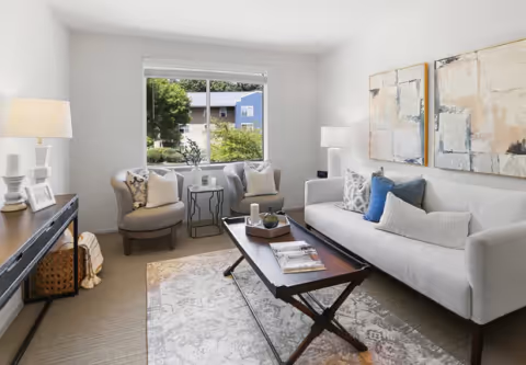 Bright furnished living room with a sofa, two armchairs, a coffee table, console table with lamp, artwork on the wall, and a window overlooking greenery.
