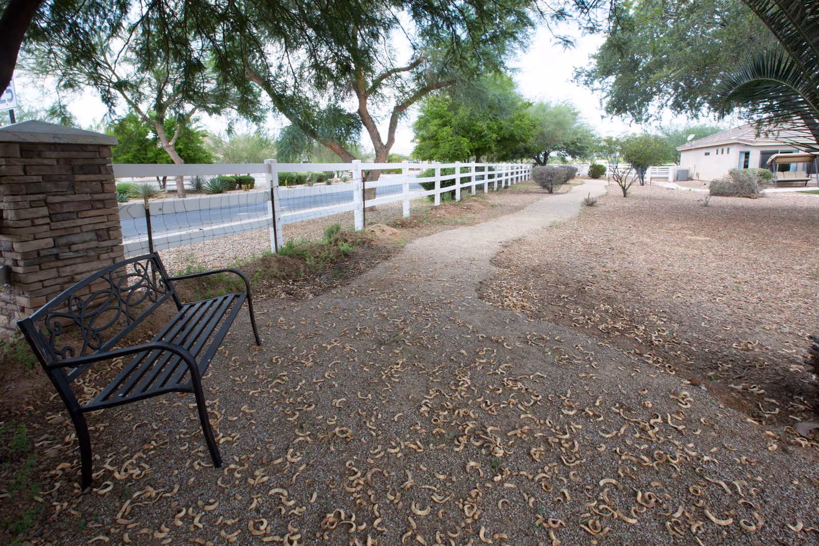 A shaded outdoor walking path lined with trees and bushes, a black metal bench on the left side near a stone pillar, and a white fence separating the path from a road. A building with a swing is visible in the background on the right.