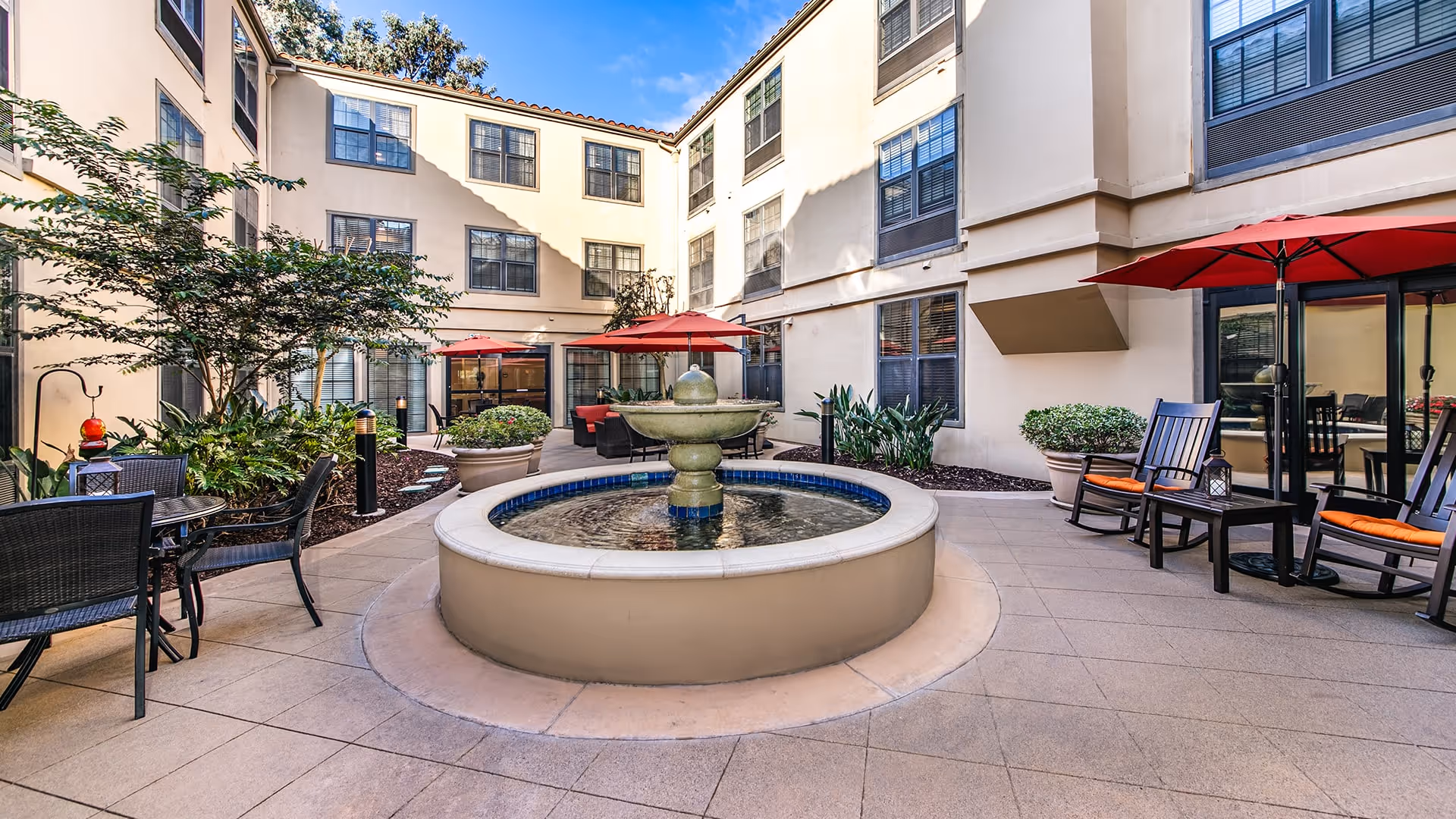 Outdoor courtyard area with a central round water fountain surrounded by tiled flooring. There are several seating arrangements including black wicker chairs and tables, rocking chairs with orange cushions, and red umbrellas providing shade. The courtyard is enclosed by a three-story building with multiple windows and some potted plants and greenery along the edges.