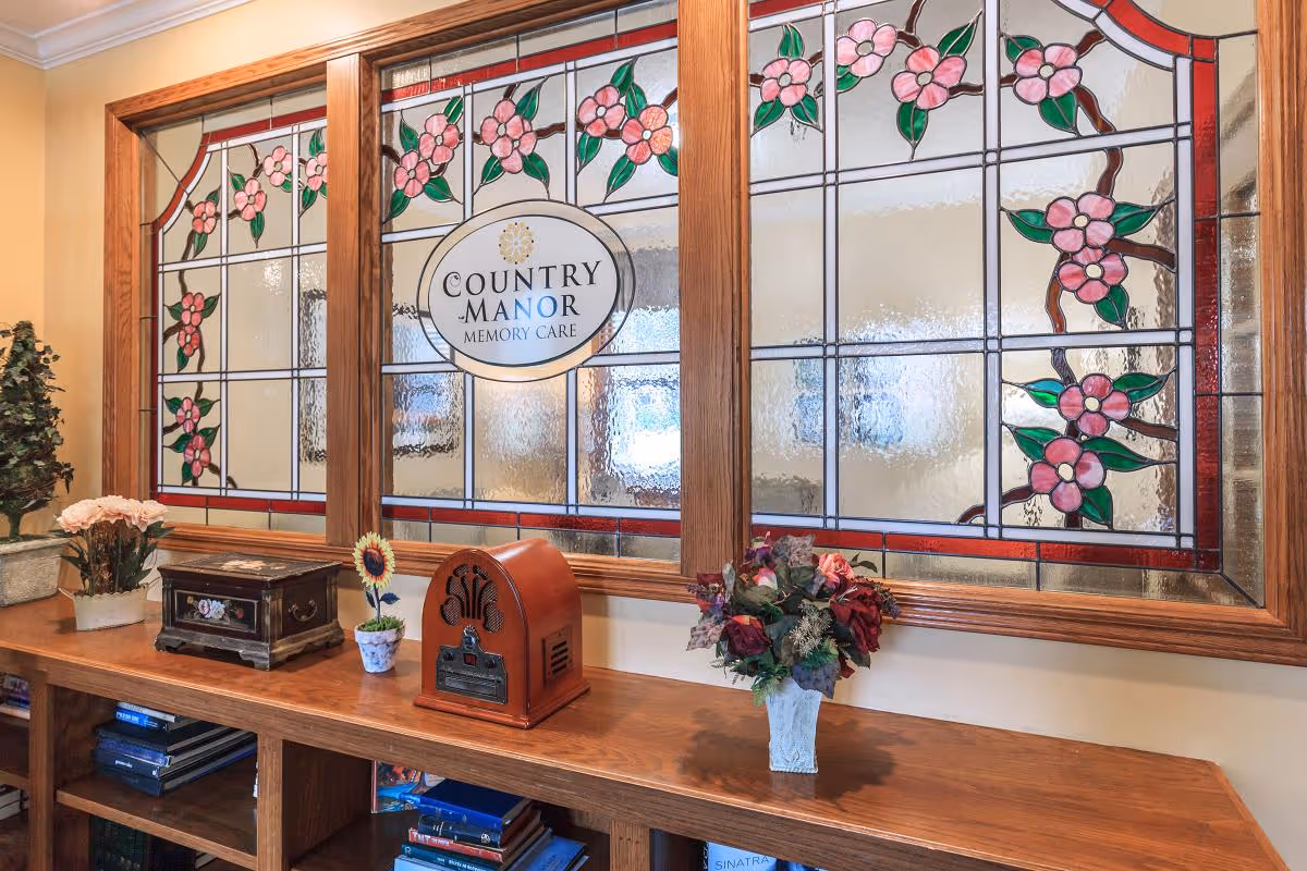 Interior view of a room with a wooden shelf holding decorative items including a vintage radio, flower pots, and a small chest. Behind the shelf is a large stained glass window with pink flowers and green leaves, featuring the text 'Country Manor Memory Care' in the center.