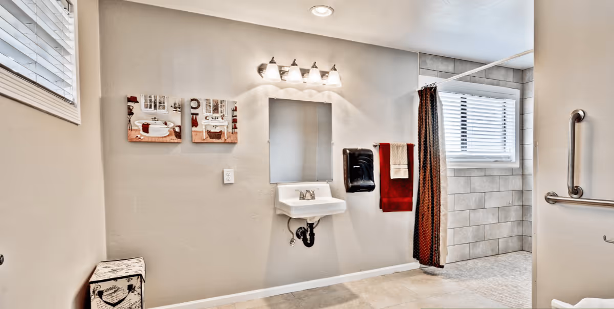 A clean and accessible bathroom with a wall-mounted sink, a mirror above it, and a light fixture with four bulbs. There are two decorative pictures on the wall, a window with blinds, a towel rack with red and beige towels, a black soap dispenser, and a walk-in tiled shower area with a grab bar.