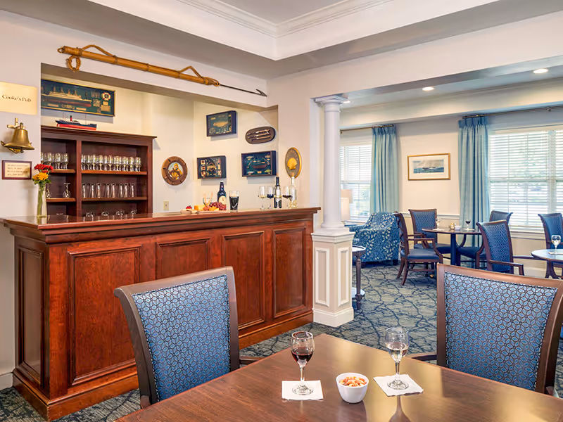 Interior view of a cozy pub-style room with a wooden bar counter, decorated with nautical-themed items on the wall. There are tables and chairs with blue patterned upholstery, glasses of wine, and a bowl of snacks on the table. The room has large windows with blue curtains allowing natural light to enter.