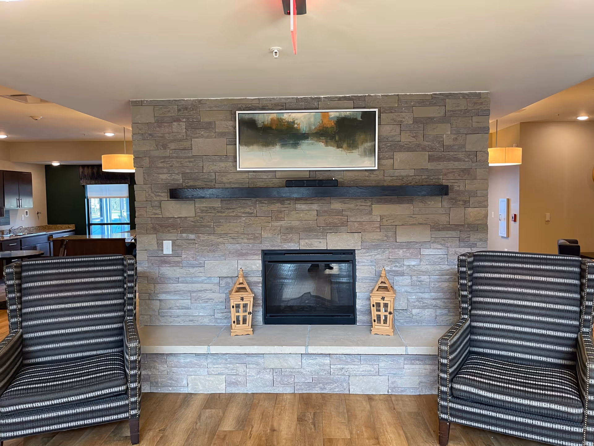 A cozy interior living area featuring a stone fireplace with a black mantel and two wooden lanterns on the hearth. Above the mantel hangs an abstract painting with muted colors. On either side of the fireplace are two striped upholstered armchairs. The room has wood flooring and warm lighting, with a glimpse of a kitchen area in the background.