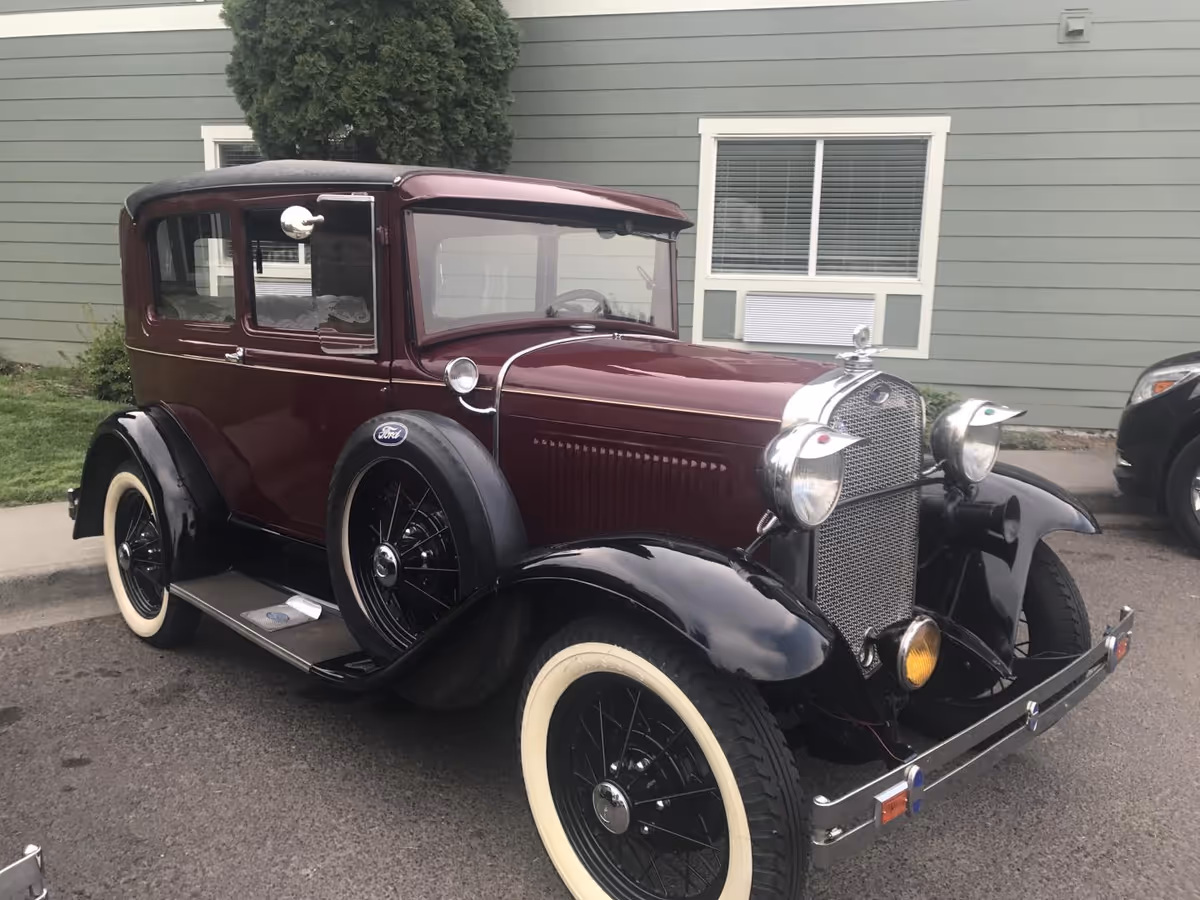 Maroon and black vintage Ford car with whitewall tires parked on a street in front of a light green building with windows.