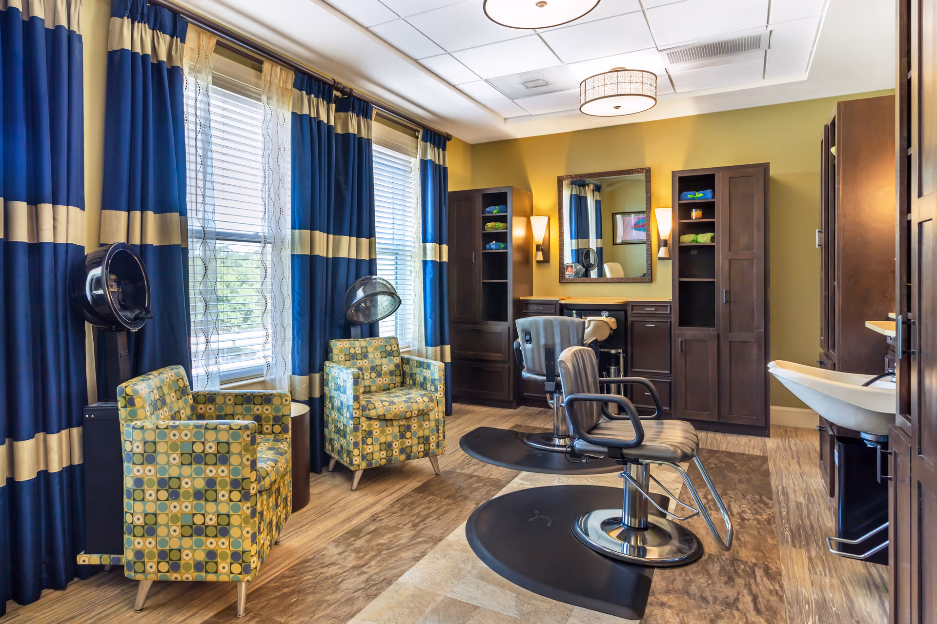 A bright salon-style interior with barber chairs, hooded dryers, patterned armchairs, sinks and dark wood cabinetry.
