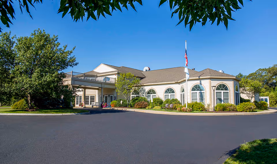 Exterior view of a single-story senior living facility building with beige siding, multiple arched windows, an American flag on a flagpole, and surrounding greenery including trees and bushes under a clear blue sky.