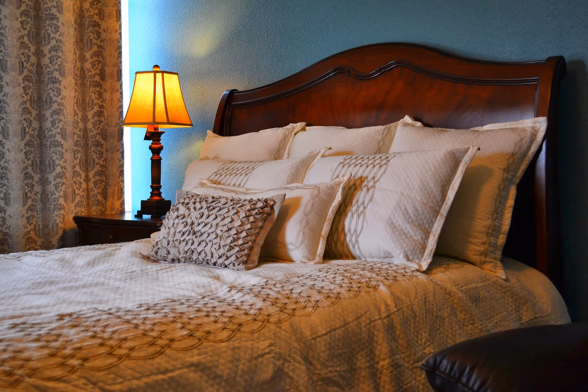 A neatly made bed with multiple decorative pillows and a wooden headboard. To the left of the bed is a nightstand with a lit table lamp and patterned curtains in the background.