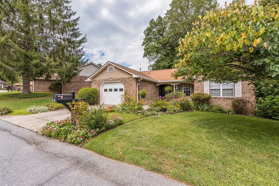 A single-story brick house with a garage, surrounded by a well-maintained lawn and colorful flower beds. There is a black mailbox near the driveway and large trees in the background under a partly cloudy sky.