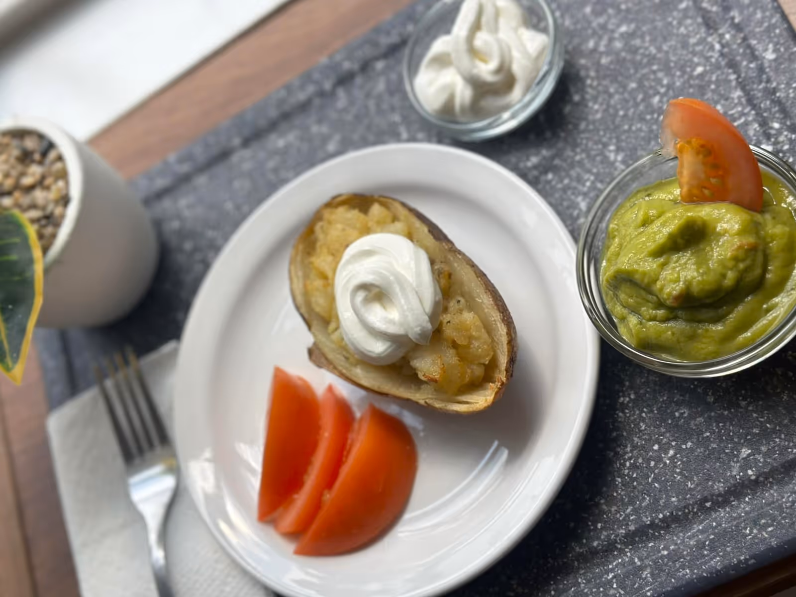 A plate with a twice-baked potato topped with sour cream, three slices of tomato, a small bowl of guacamole garnished with a tomato slice, and a small bowl of sour cream on a tray next to a fork and a potted plant.