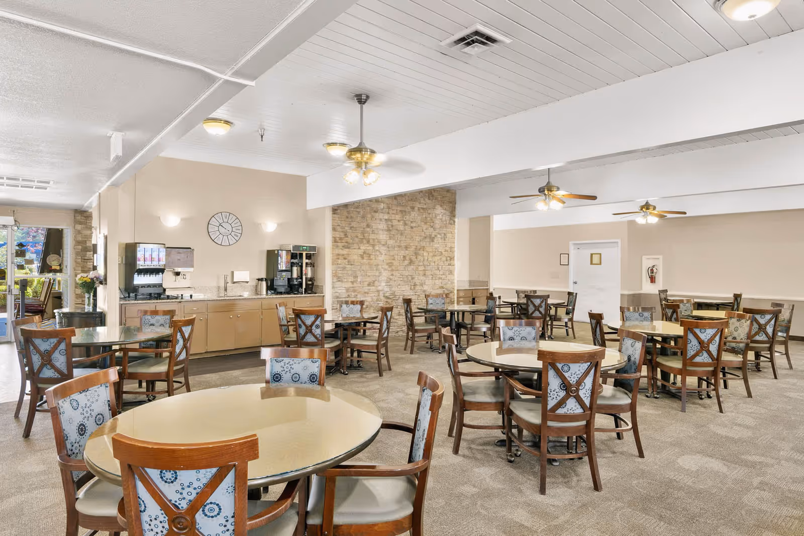 A spacious dining room with multiple round tables and wooden chairs featuring patterned cushions. The room has beige walls, a stone accent wall, ceiling fans with lights, and a beverage station with coffee and soda machines. Large windows allow natural light to enter the room.