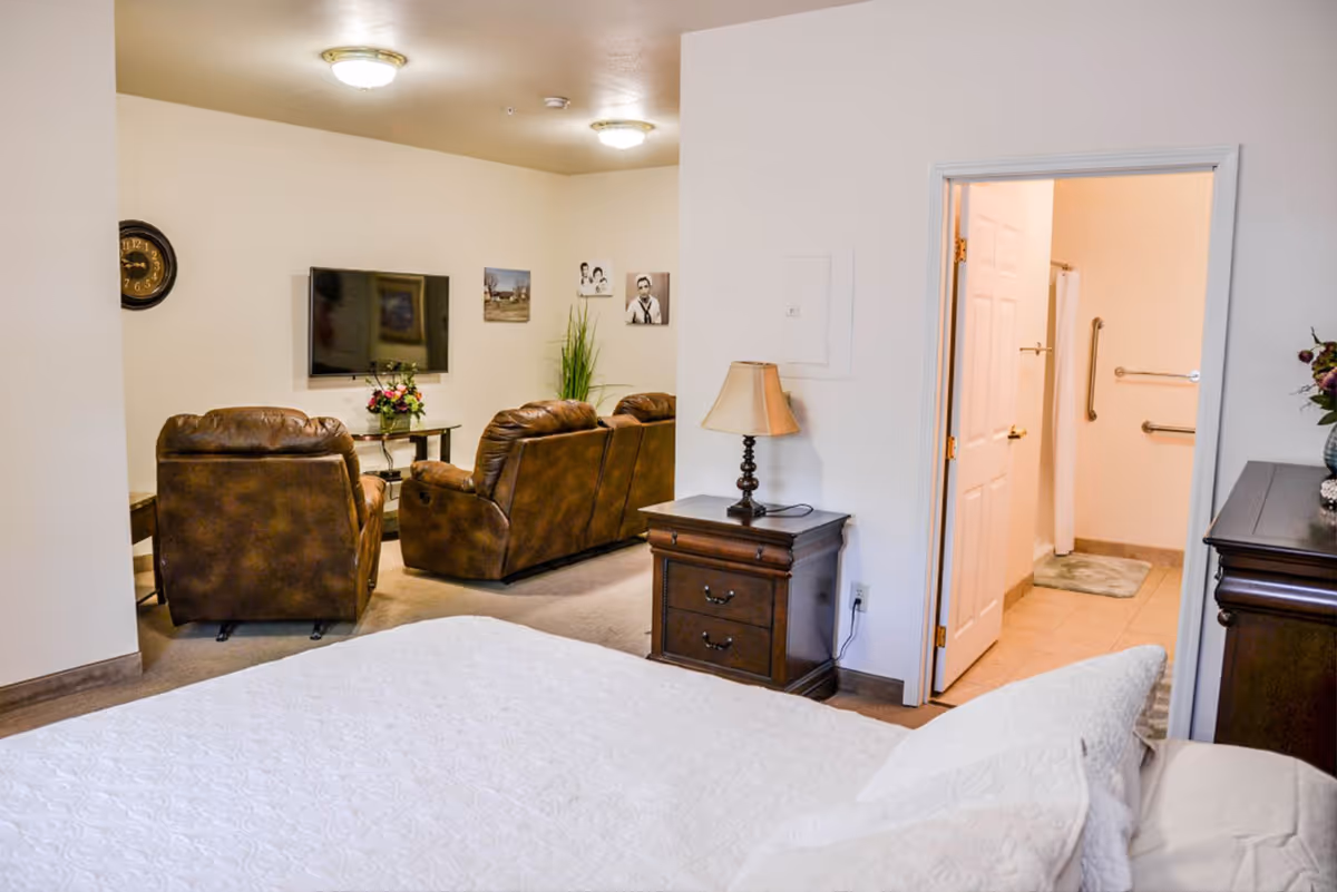 A senior living facility room featuring a bed with white bedding in the foreground, a wooden nightstand with a lamp, and an open door leading to a bathroom with grab bars. In the background, there is a sitting area with brown leather chairs, a wall-mounted TV, a clock, and framed pictures on the wall.