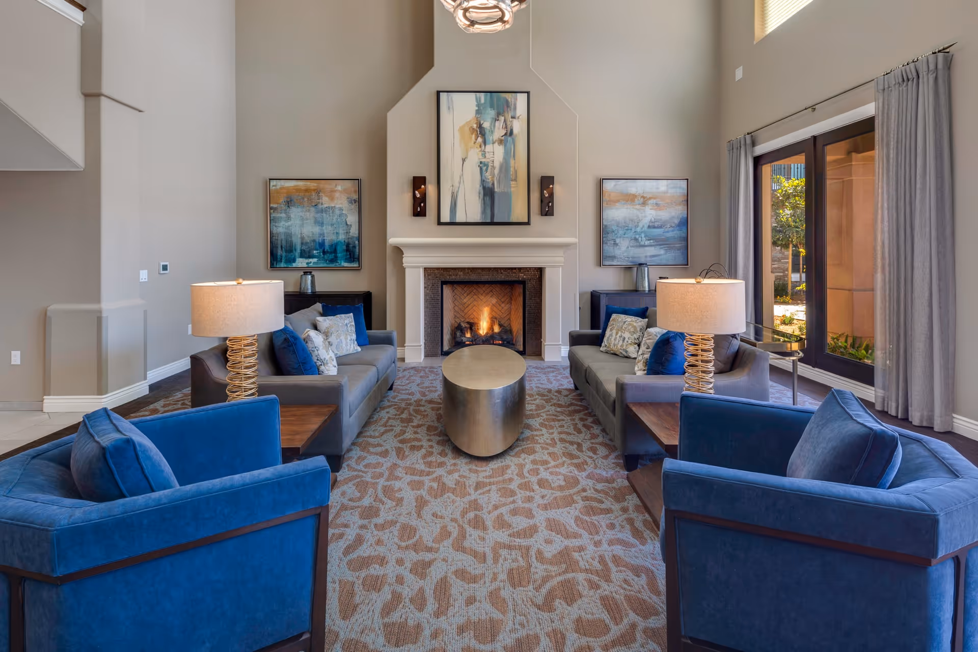 A spacious and well-lit living room with two gray sofas and two blue armchairs arranged around a modern oval coffee table. The room features a lit fireplace with abstract artwork above it and two additional paintings on either side. There are two table lamps with beige shades on wooden side tables next to the sofas. Large windows with gray curtains allow natural light to enter the room.