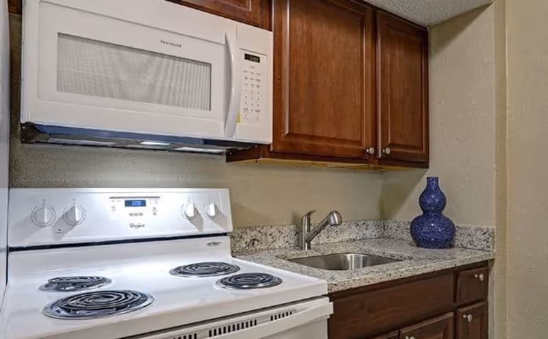 A kitchen area featuring a white electric stove with four coil burners and a white microwave mounted above it. There are wooden cabinets above and below a granite countertop with a small stainless steel sink and a blue decorative vase on the right side.