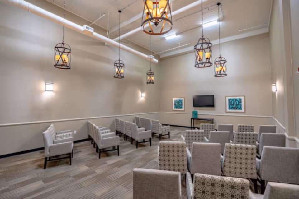 A small conference or meeting room with multiple rows of beige and patterned chairs facing a wall-mounted flat screen TV. The room has beige walls, modern hanging lantern-style light fixtures, and two framed artworks on the wall beside the TV.