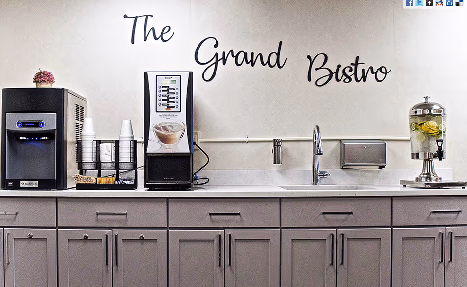 A countertop with a coffee machine, cups, a water dispenser with lemon slices, a sink, and cabinets below. The wall behind has the words 'The Grand Bistro' written in decorative script.