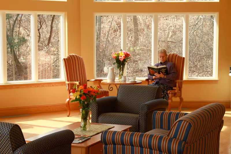 A cozy living room area with large windows showing a view of leafless trees outside. The room has comfortable upholstered armchairs and a wooden coffee table with a vase of flowers. An elderly woman is sitting in one of the armchairs near the windows, reading a book.
