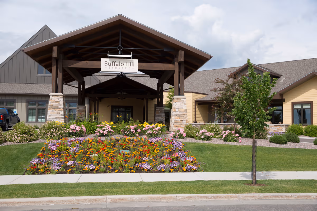 Front exterior view of Immanuel Living at Buffalo Hill facility with a covered entrance supported by stone pillars, a sign reading 'Buffalo Hill Terrace,' colorful flower beds, green lawn, and a young tree in the foreground under a partly cloudy sky.