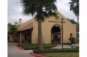 Exterior view of a nursing center building with a beige stucco facade, a peaked roof, and an arched entrance. There are palm trees and shrubs in front of the building, and a red curb along the driveway.