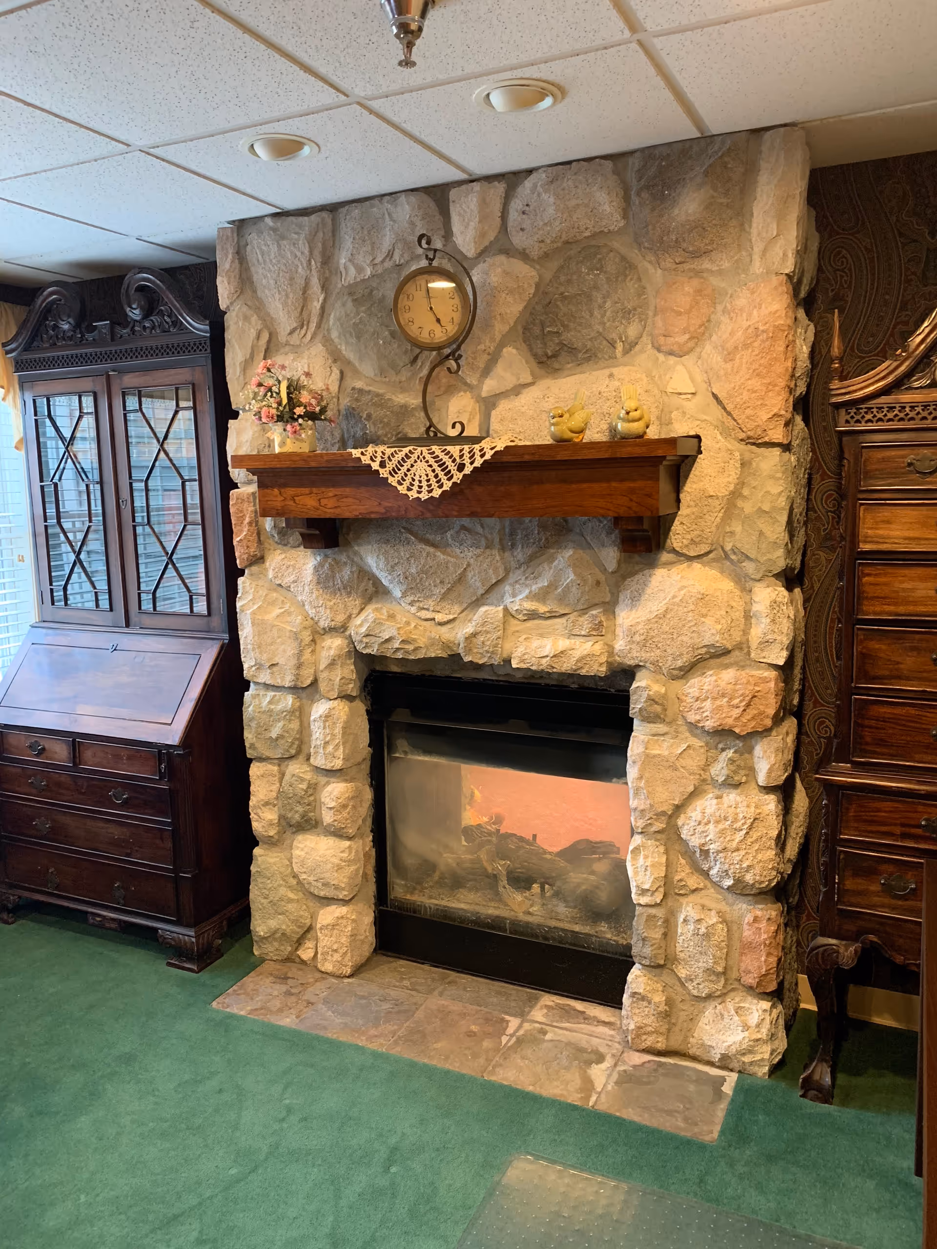 A cozy living room area featuring a stone fireplace with a wooden mantel. On the mantel, there is a decorative clock, a small vase with pink flowers, and two yellow bird figurines. To the left of the fireplace is a dark wooden cabinet with glass doors and a drop-front desk. The floor is covered with green carpet.