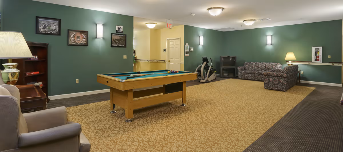 A recreational room with a pool table in the center, an exercise bike near the back wall, a floral patterned couch, an armchair, a TV on a stand, and framed pictures on green walls. The room has carpeted flooring with a patterned design and is well-lit with ceiling and wall lights.