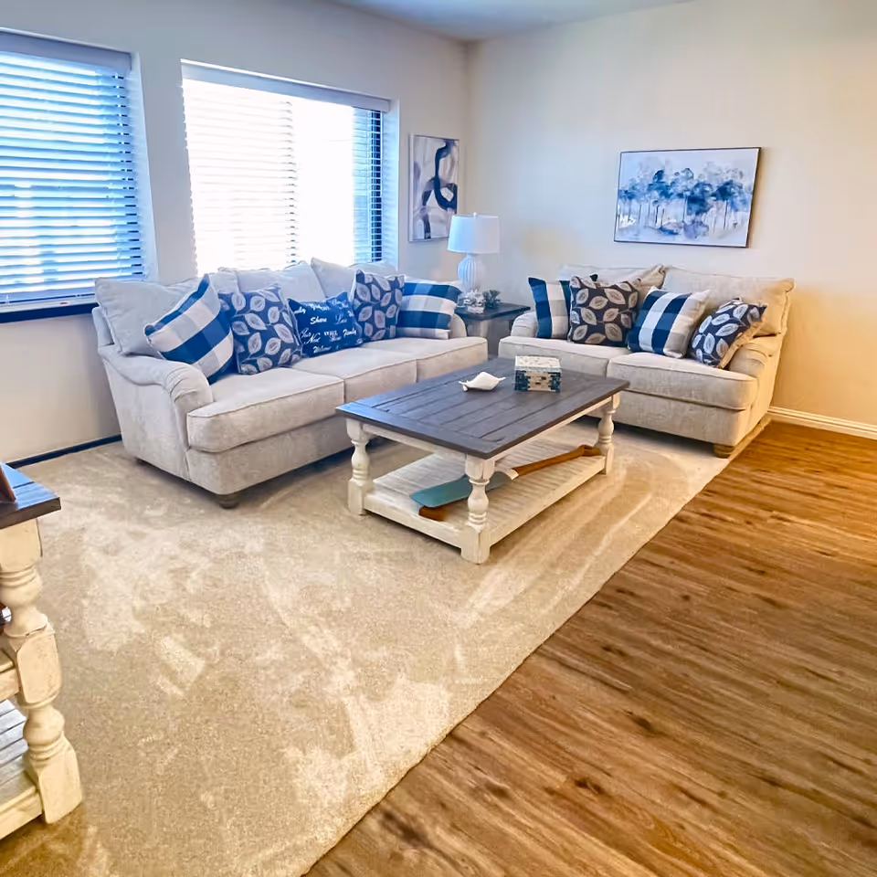 A bright living room with two beige sofas adorned with blue and white patterned cushions, a wooden coffee table with a decorative box and a small dish, a side table with a white lamp, and two windows with blinds letting in natural light. The room has a beige carpet and wooden flooring, with abstract artwork on the walls.
