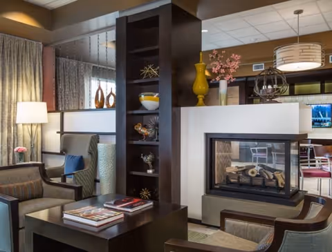 A cozy living room area featuring a modern double-sided fireplace, comfortable armchairs, a coffee table with books, decorative shelves with vases and ornaments, and soft lighting from a floor lamp and ceiling fixture.