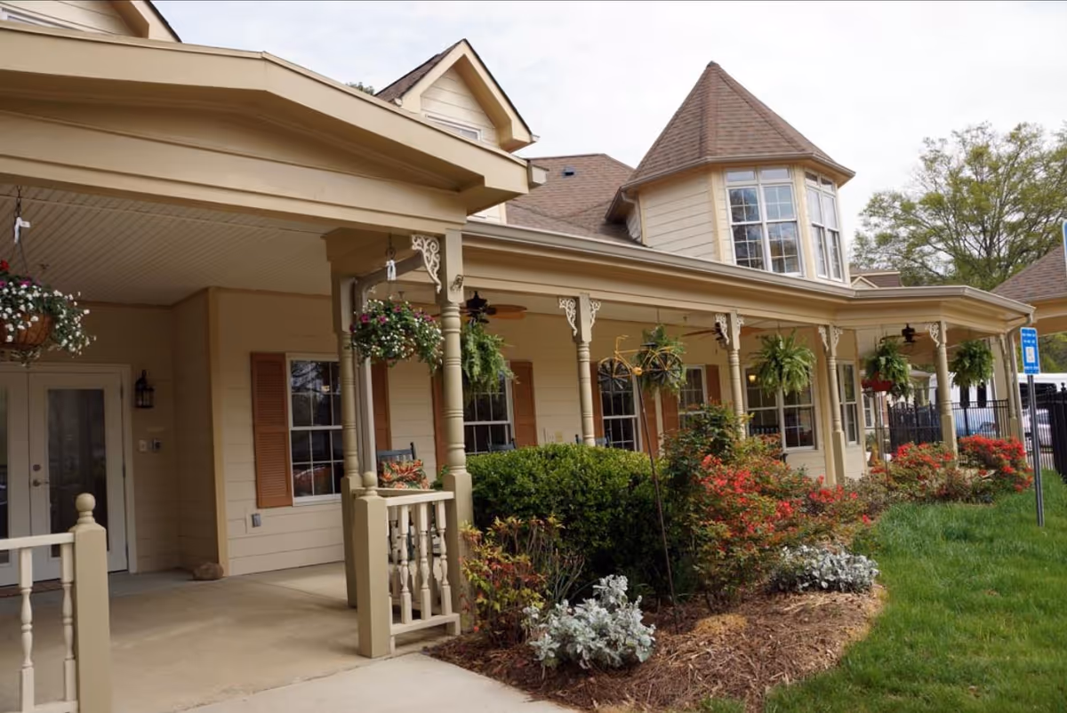 Front exterior of a beige senior living building with a covered porch, hanging plants, and landscaped shrubs.