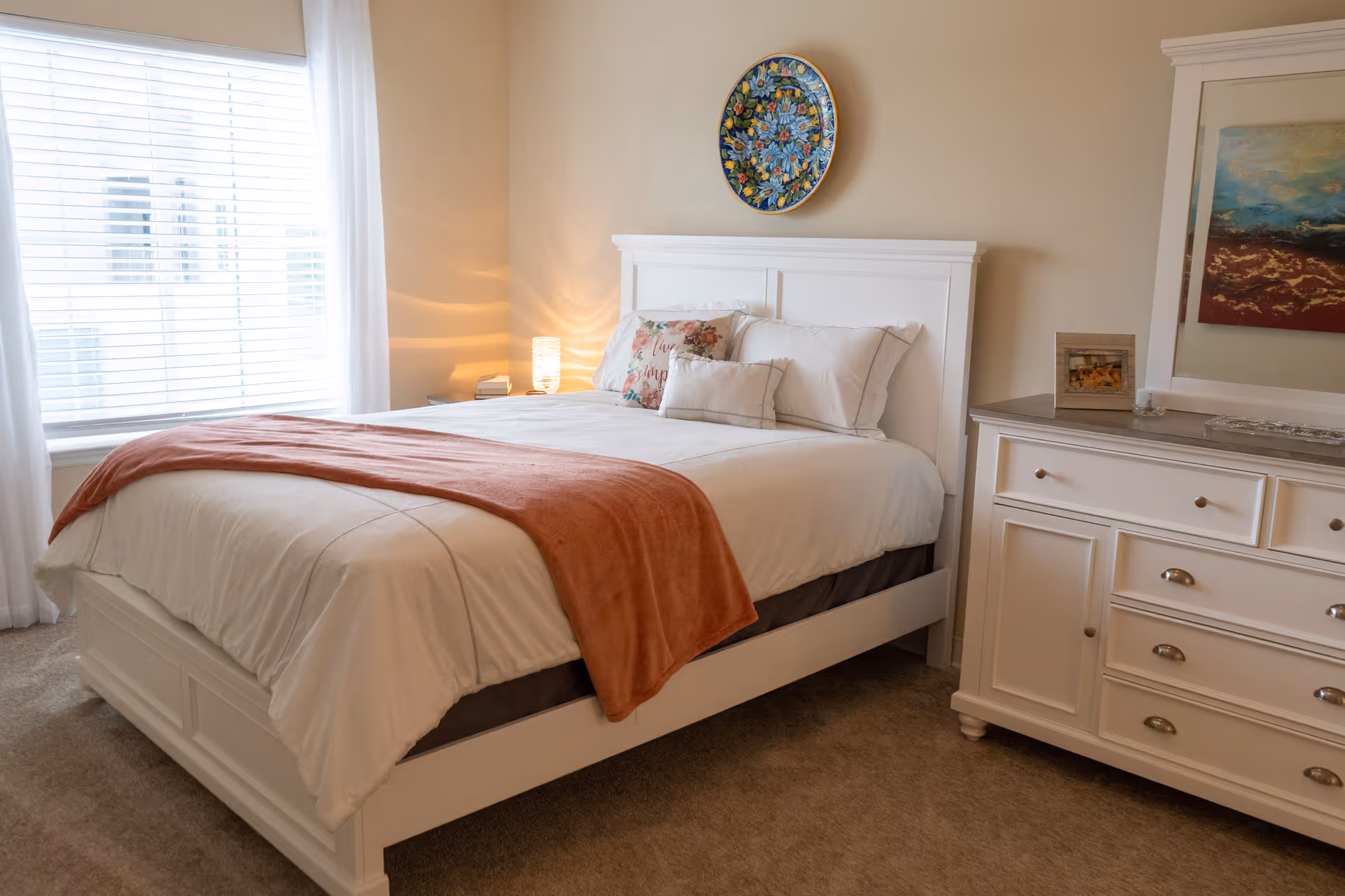A cozy bedroom with a white wooden bed frame and a neatly made bed featuring white bedding and a rust-colored throw blanket. There are decorative pillows on the bed, a small bedside table with a lamp, and a white dresser with a framed picture and a mirror above it. A colorful decorative plate hangs on the wall above the bed. Natural light comes through a window with white blinds and curtains.