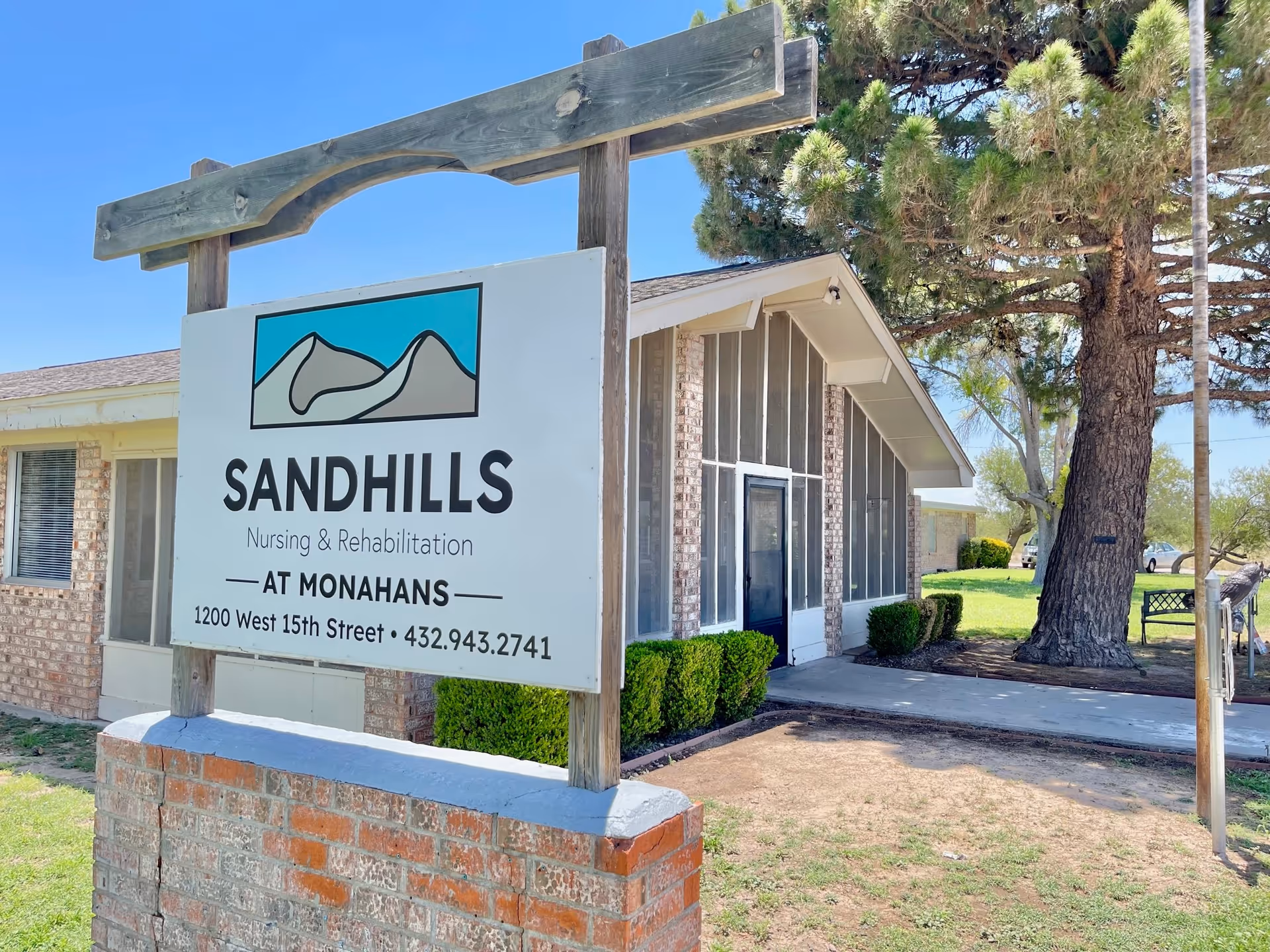 Outdoor view of Sandhills Nursing & Rehabilitation facility sign at Monahans, showing the building entrance with large windows, a tree, and a bench in the background under a clear blue sky.
