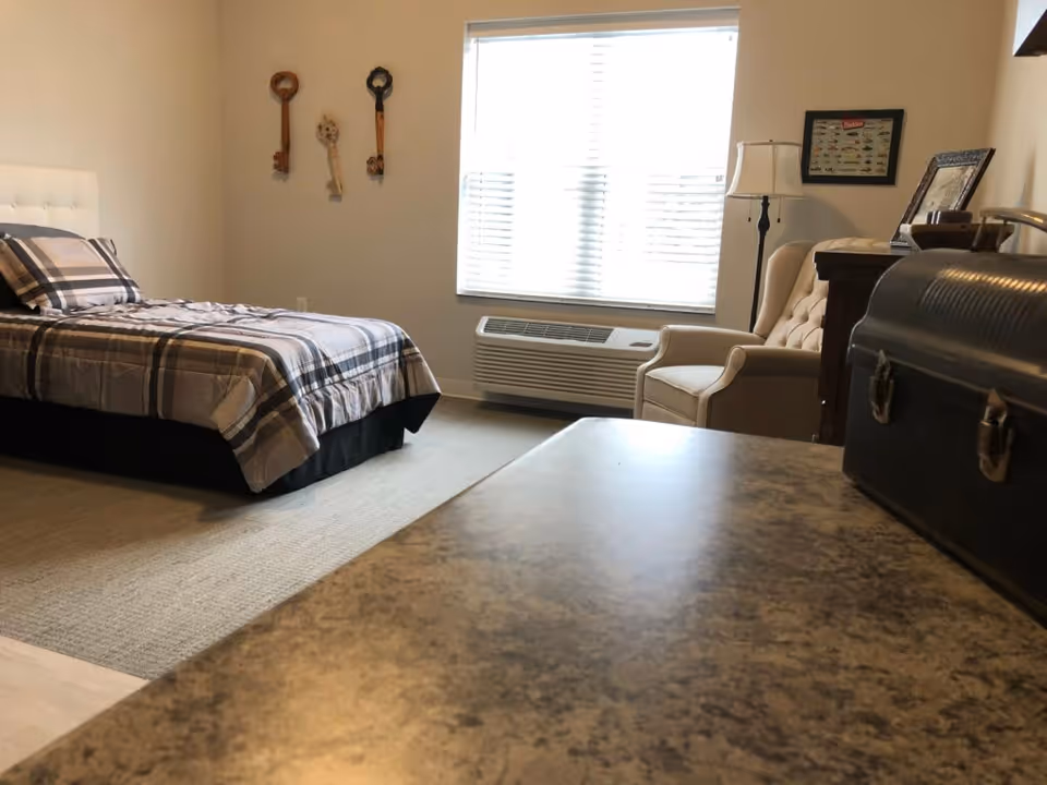 A furnished bedroom with a single bed in plaid bedding, an armchair, dresser, and a large window with blinds.