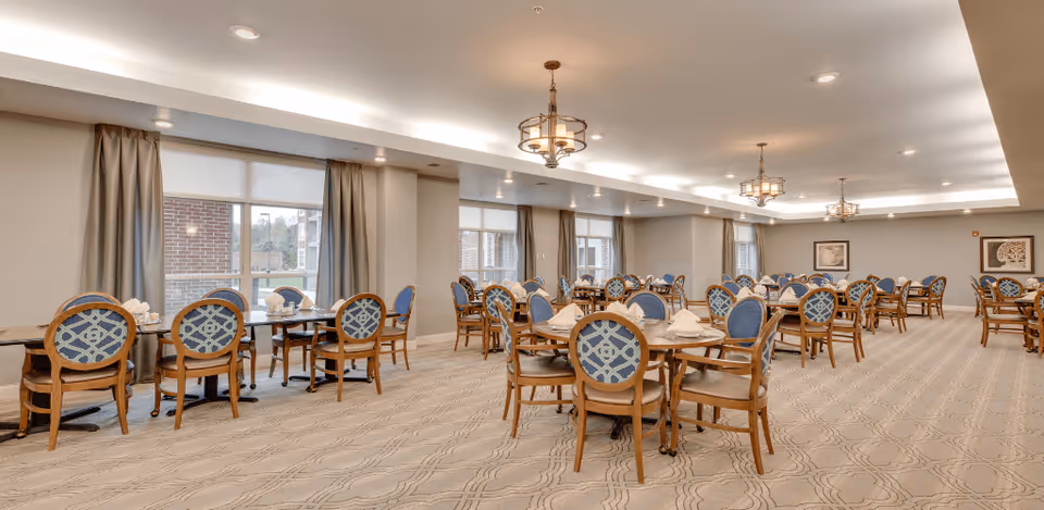 A spacious dining room with multiple round and rectangular tables, each surrounded by wooden chairs with blue patterned cushions. The room has large windows with beige curtains, soft overhead lighting, and framed artwork on the walls. The carpet has a subtle geometric pattern.