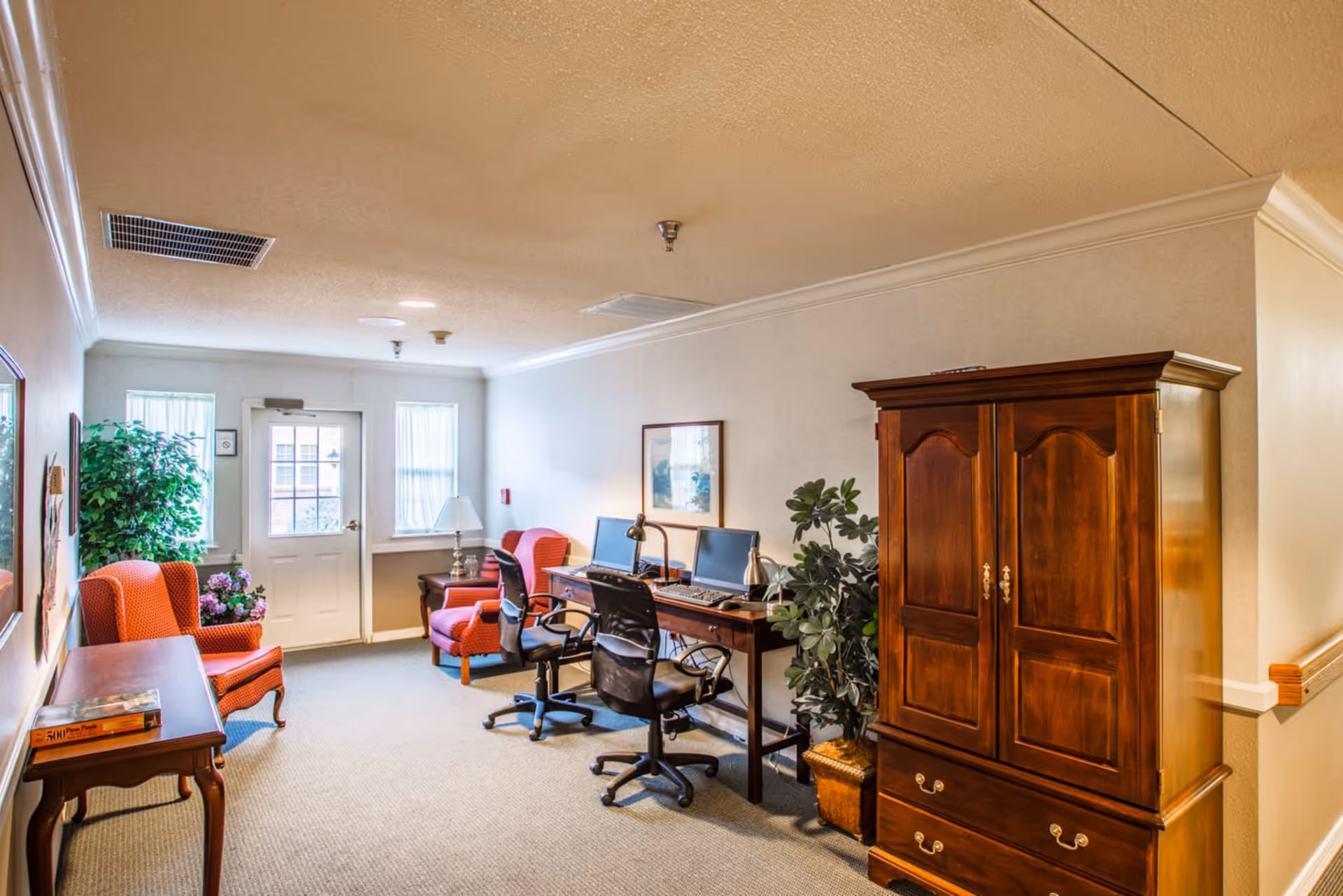 A well-lit interior room with a wooden armoire on the right, two office chairs at a desk with two computer monitors, and two orange upholstered armchairs near a door with windows. There are plants and framed artwork on the walls, creating a cozy and functional space.