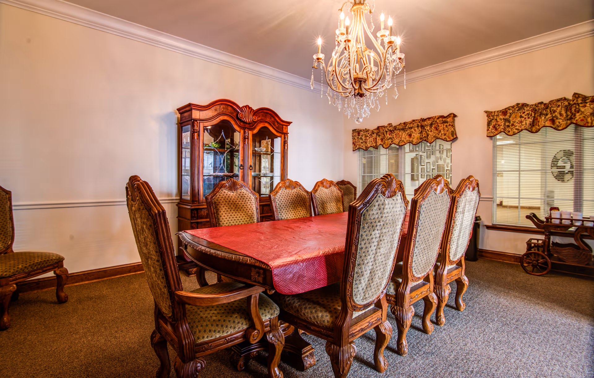 A formal dining room with a long wooden table covered by a red tablecloth, surrounded by eight ornate wooden chairs with upholstered seats and backs. A wooden china cabinet with glass doors is against the wall, and a crystal chandelier hangs from the ceiling. The room has carpeted flooring, white walls with molding, and windows with floral valances.
