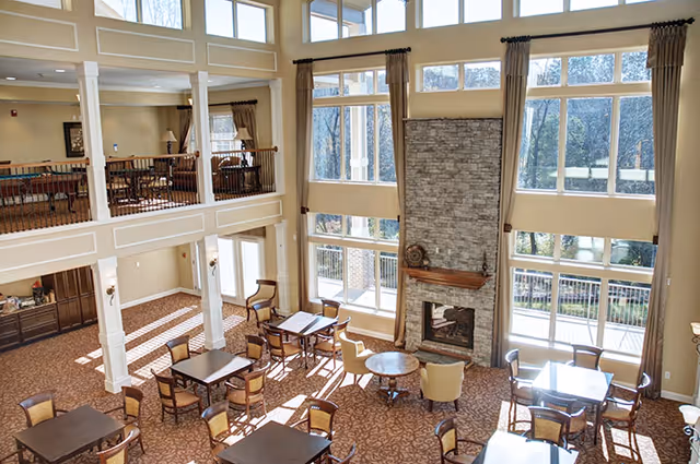 Spacious two-story common area with large windows letting in natural light, multiple tables and chairs arranged for seating, a tall stone fireplace with a wooden mantel, and beige walls with decorative molding. The upper level has a railing overlooking the area below.