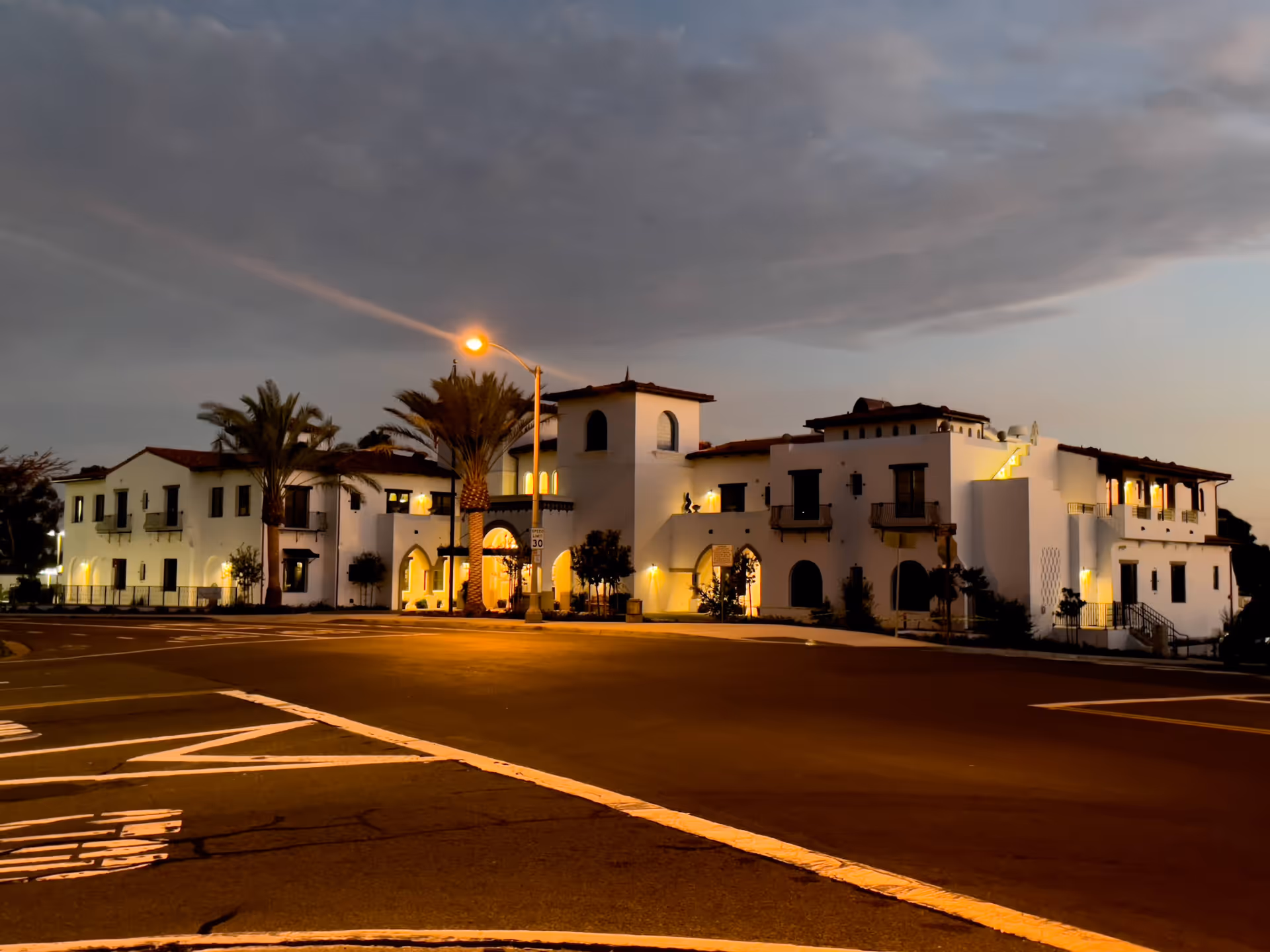 Exterior view of a large, white, Mediterranean-style building at dusk with warm lights glowing from the windows and palm trees in front. The building is situated at a street corner with visible road markings and a street lamp illuminating the scene.