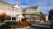Exterior view of a multi-story assisted living facility building with beige siding, white trim, and a covered entrance. There is a flagpole with an American flag in front, surrounded by landscaped bushes and trees with autumn foliage. The sky is clear and blue.