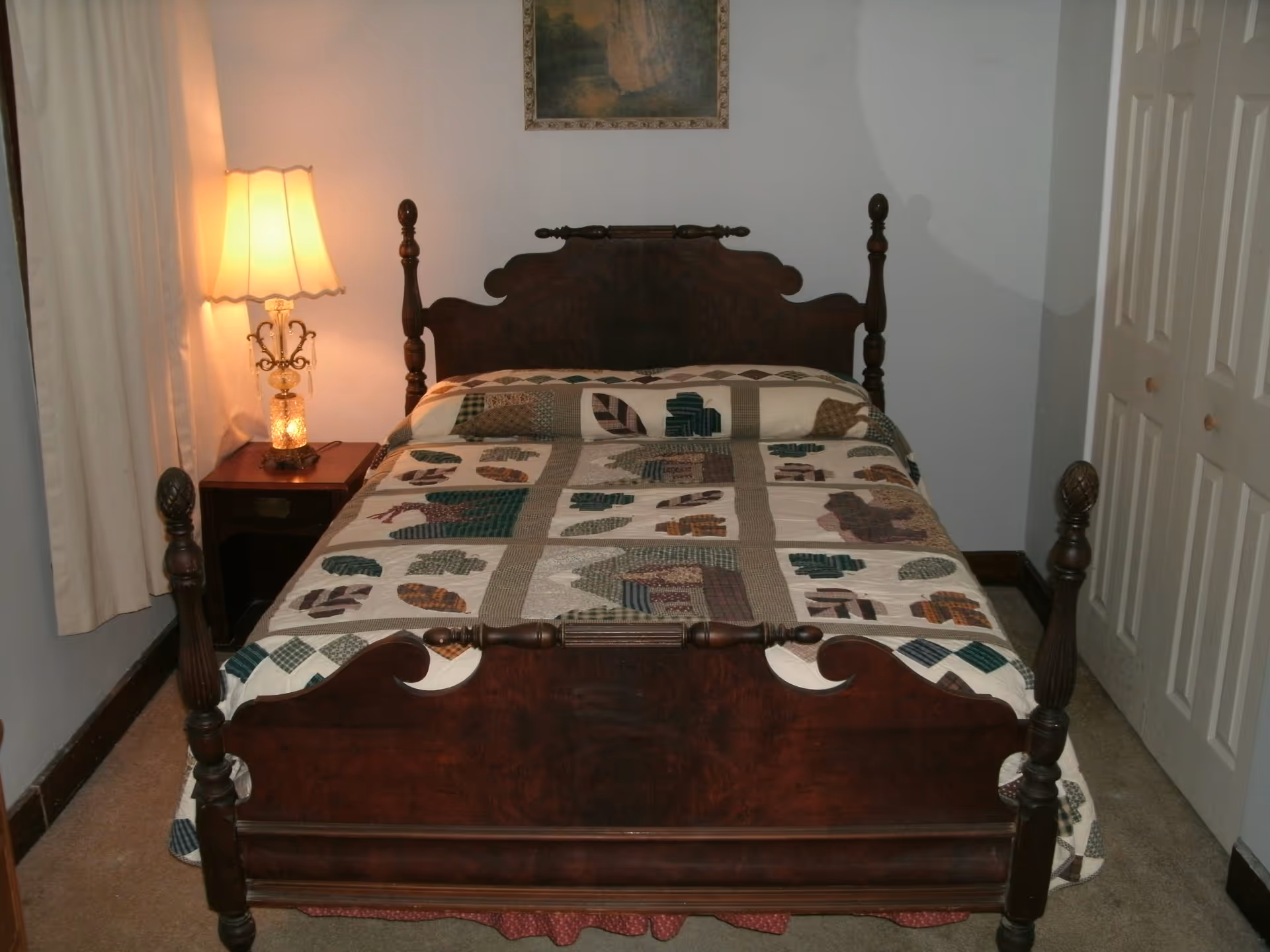 Antique wooden bed with a patchwork quilt, nightstand and lamp in a small bedroom.