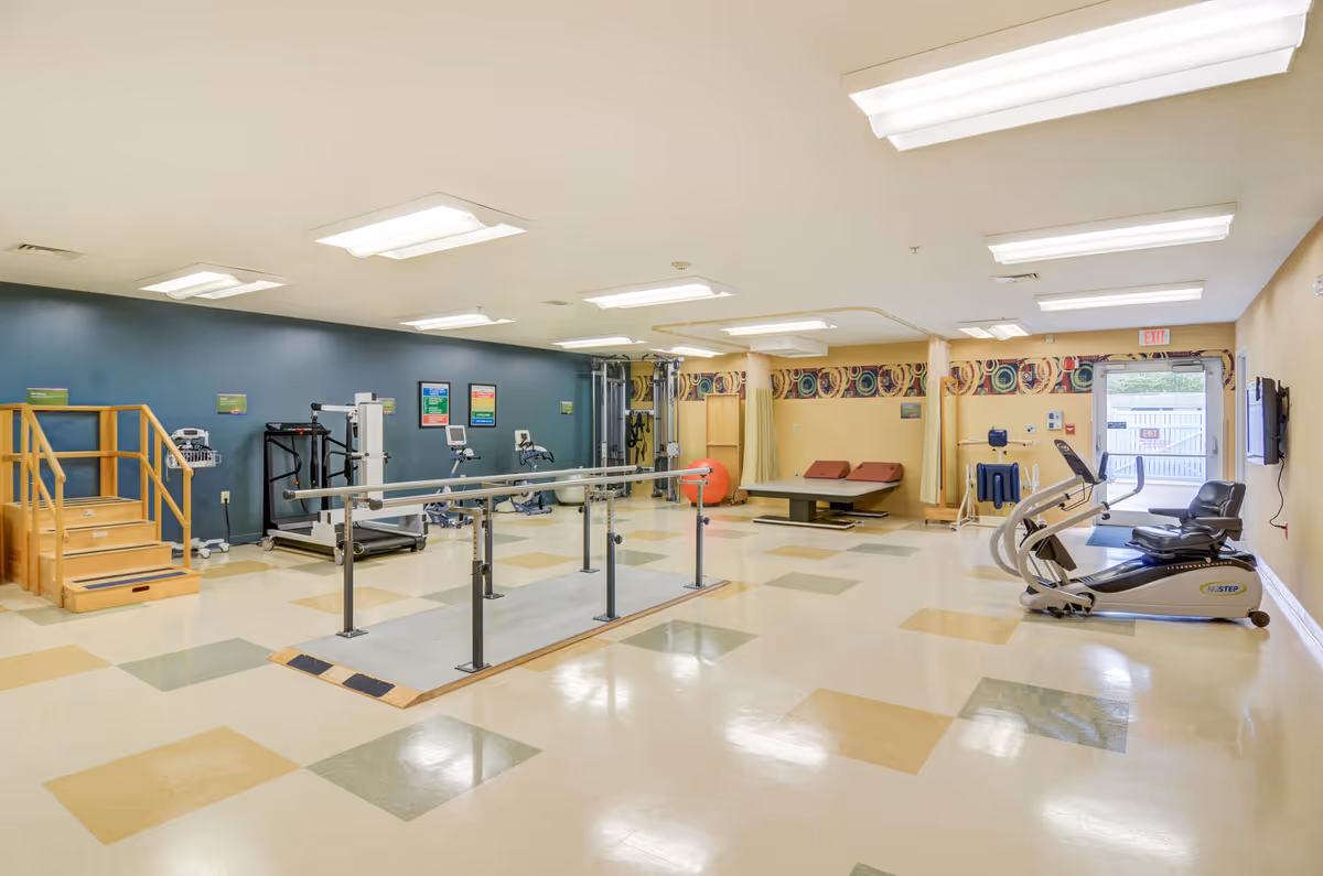 A spacious physical therapy or rehabilitation room with exercise equipment including parallel bars, a treadmill, stationary bikes, therapy steps, and treatment tables. The room has a patterned floor with beige, green, and yellow tiles, light yellow walls, and a blue accent wall with posters. There is an exit door with a window letting in natural light.