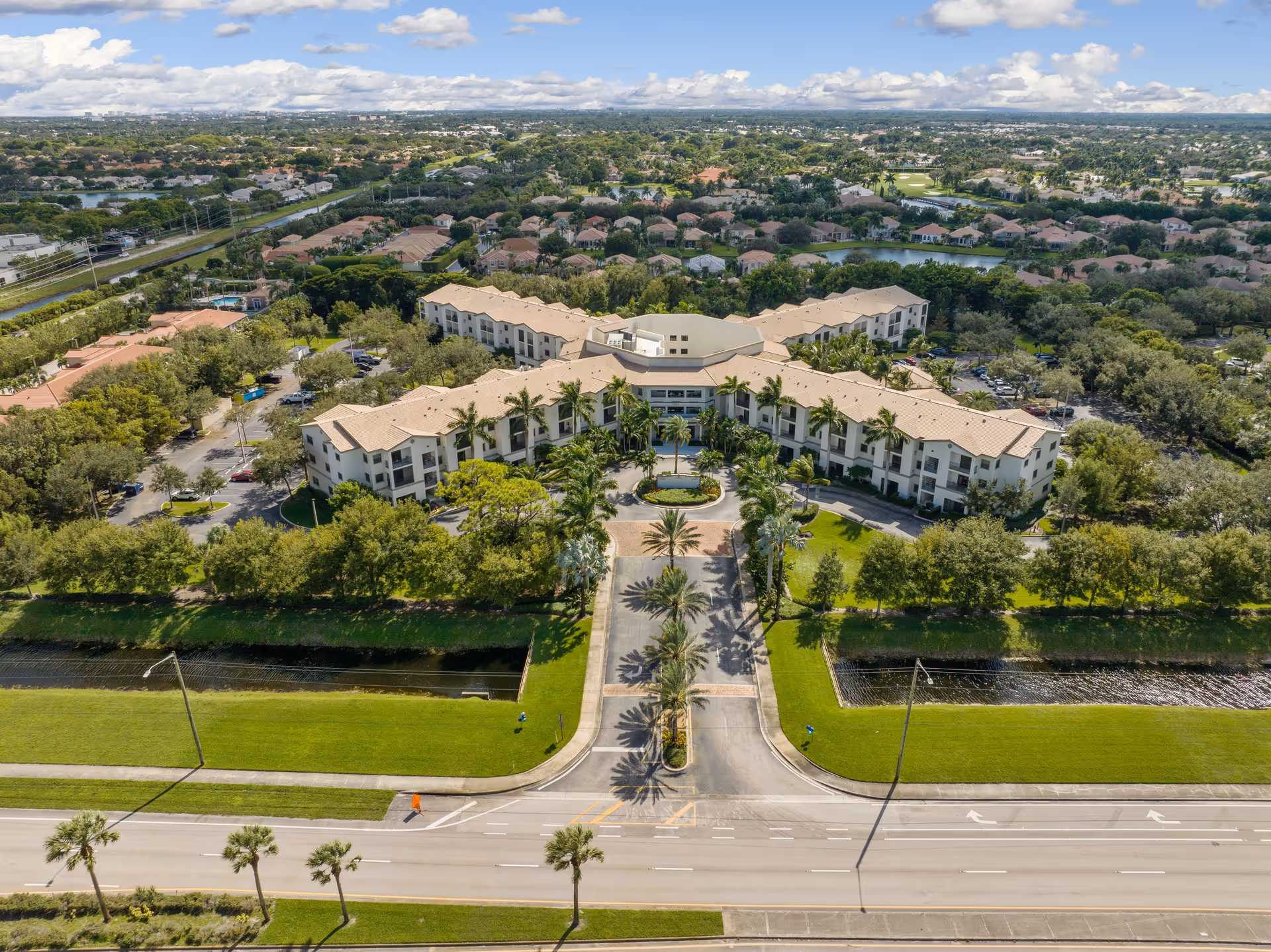 Aerial view of a large senior living facility named Verena at Delray, surrounded by lush greenery, palm trees, and a well-maintained lawn. The building has a beige exterior with a tiled roof and is situated near a water canal. The entrance driveway is lined with palm trees and leads to a circular drop-off area in front of the building. Residential neighborhoods and a golf course are visible in the background under a partly cloudy sky.